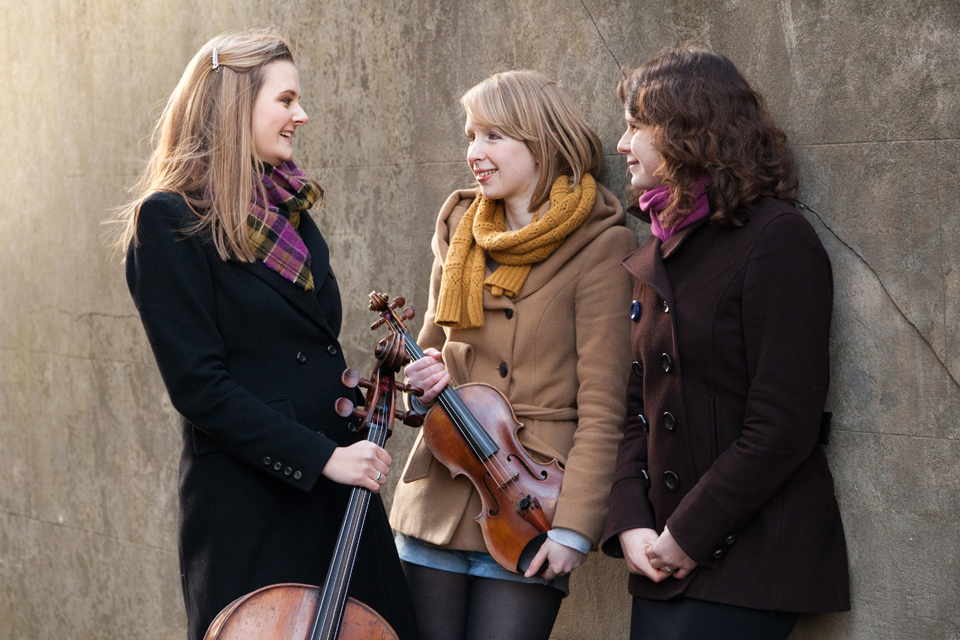 A young women, with blonde hair, holding a cello, speaking to two women, with blonde and brown hair, one of them holding a violin, standing outside.